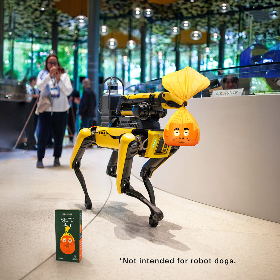 A robot dog at a trade show carrying a tied Stable Genius bag, with the carton on the floor beside it.